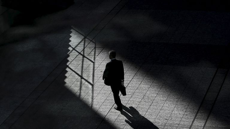 Ένας άνδρας με γραβάτα και σακίδιο περπατάει σε σκοτεινό χώρο, με το φως να φωτίζει ένα τμήμα του δαπέδου και τη σκιά του να απλώνεται μπροστά του.