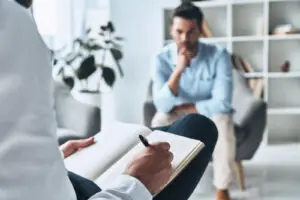 ένας θεραπευτής ή σύμβουλος γράφει σε ένα σημειωματάριο κατά τη διάρκεια μιας συνεδρίας. Στο background, υπάρχει ένας άνδρας που κάθεται στον καναπέ, φαίνεται σκεπτικός ή ανήσυχος, πιθανώς πελάτης ή ασθενής